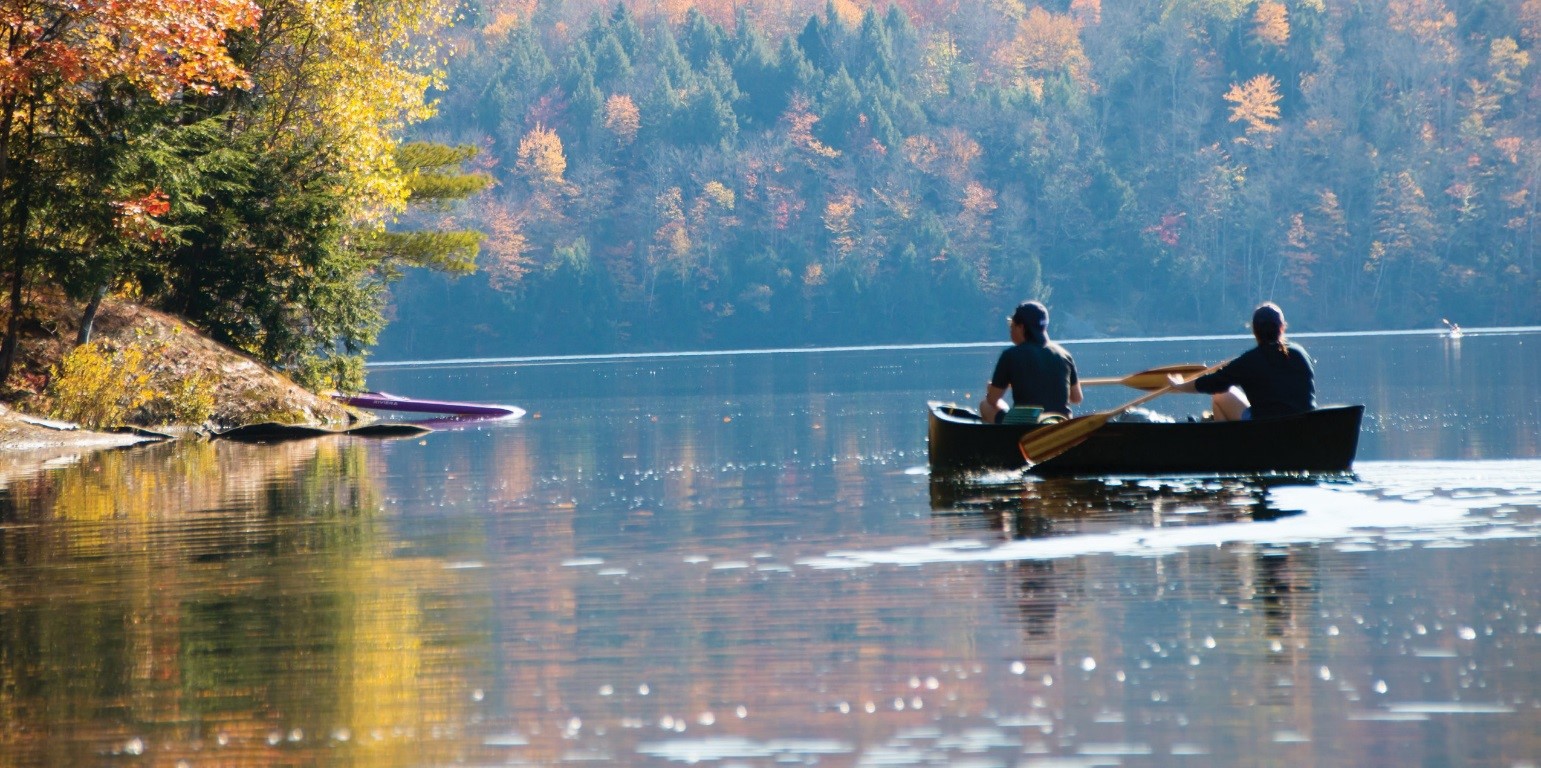 A man and woman paddle a canoe on a calm lake edged by colorful trees in fall.