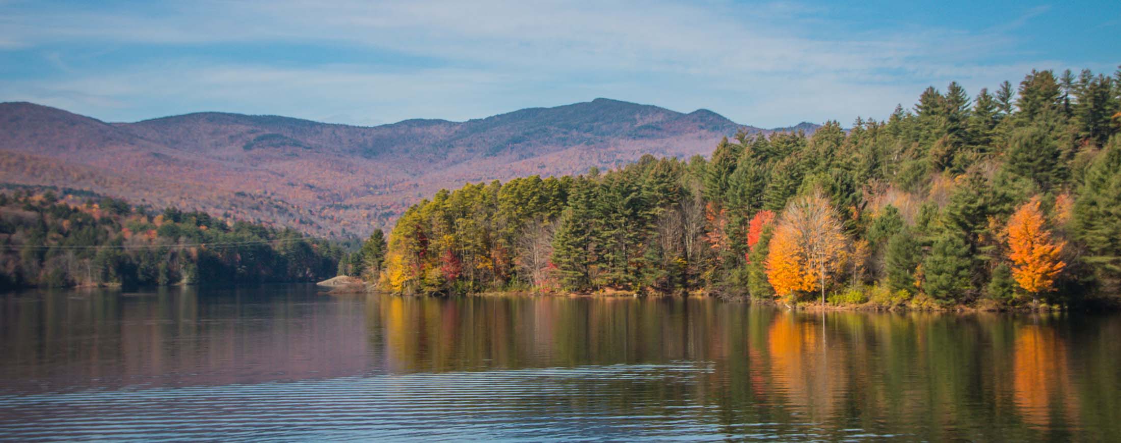 A view of a lake surrounded by mountains and trees that are turning red, orange and yellow in the fall. ge an 