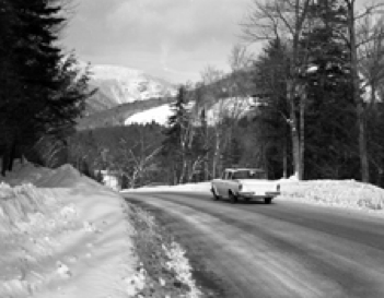 A black-and-white photo of a 1950s-style car driving through the mountains in winter. 