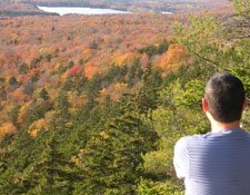 A man who is hiking takes in the view from a mountain top of fall foliage with a lake in the distance.