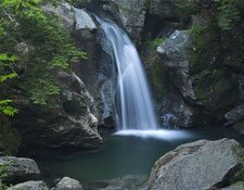 A waterfall surrounded by boulders plunges from atop a small cliff in a forest. 