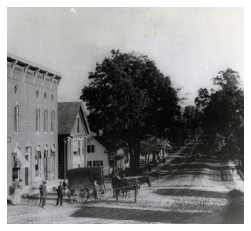 A black and white photo of a village street with horses and buggies.