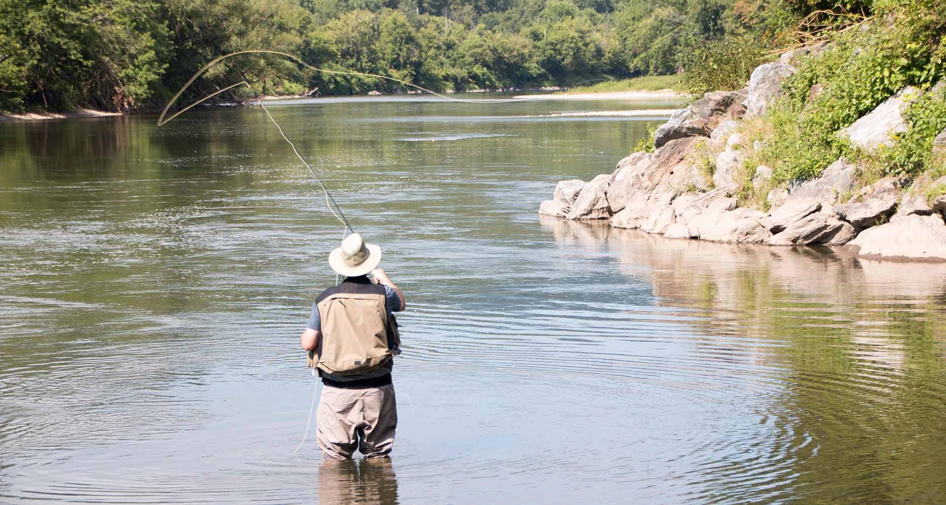 A man stands in a river wearing waders and casting a fishing pole.