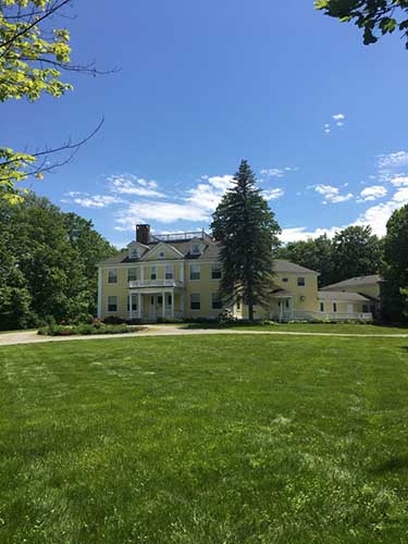 A yellow mansion with columns and a circular drive is located across a lush, green field. 