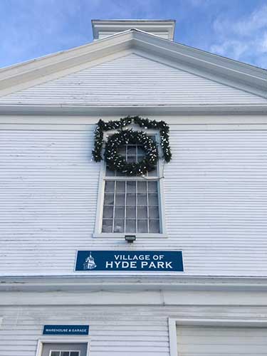 The upper frame of a white clapboard building on which the words "Village of Hyde Park" appear, above a wreath framing a window.