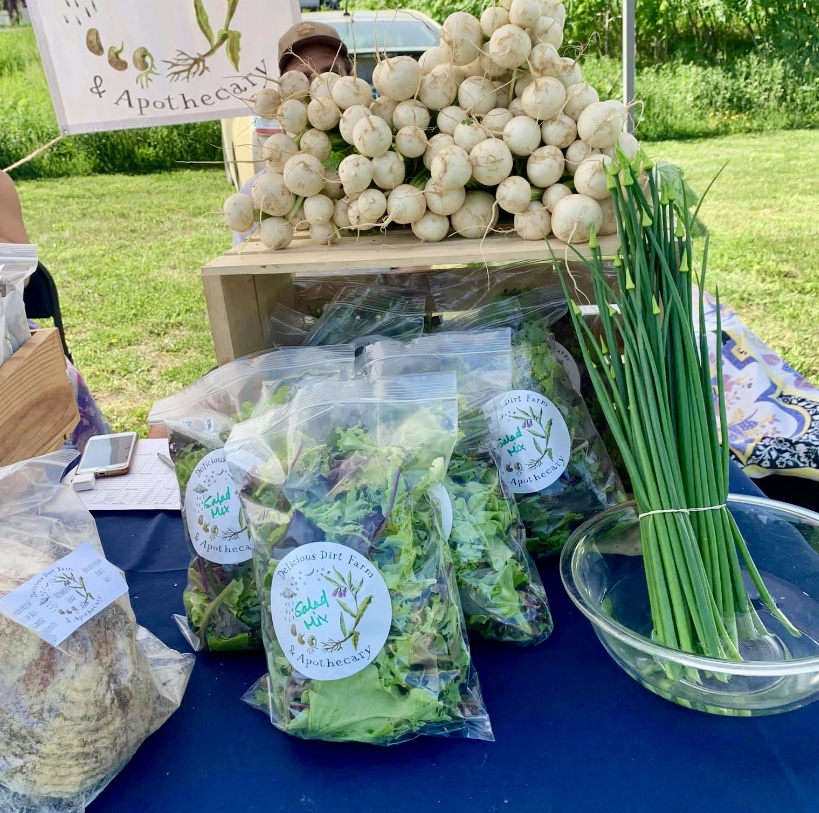 Greens, garlic scapes and turnips are laid out on a table, for sale at a summer farmers market.