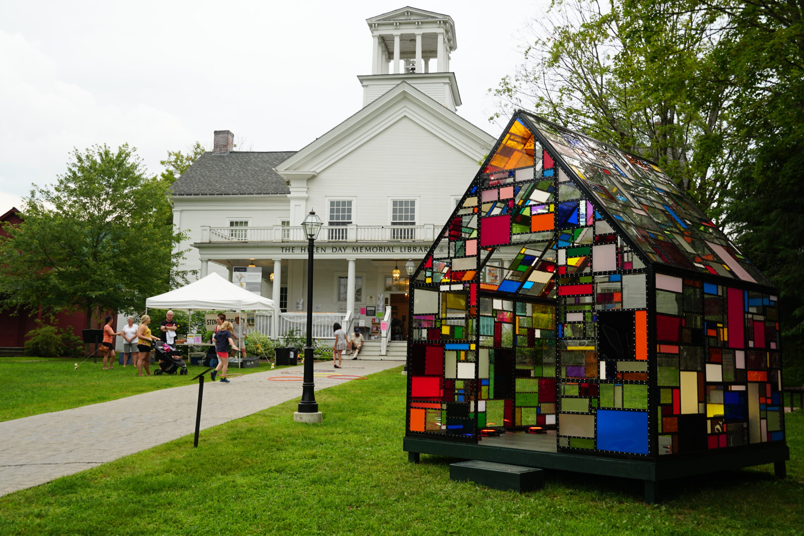 A small house made of stained glass stanks before a white library building in summer.